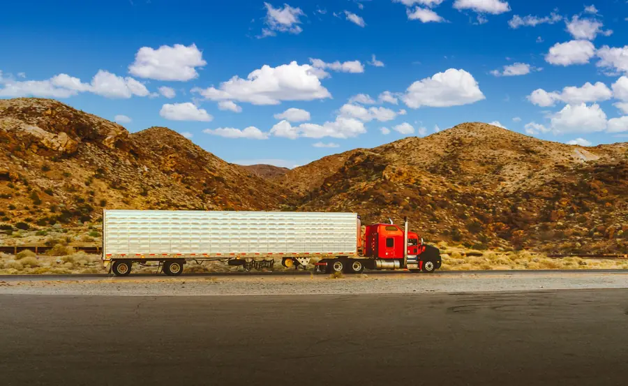 A red semi truck pulling a long trailer along a desert highway beneath rocky hills and a blue sky.