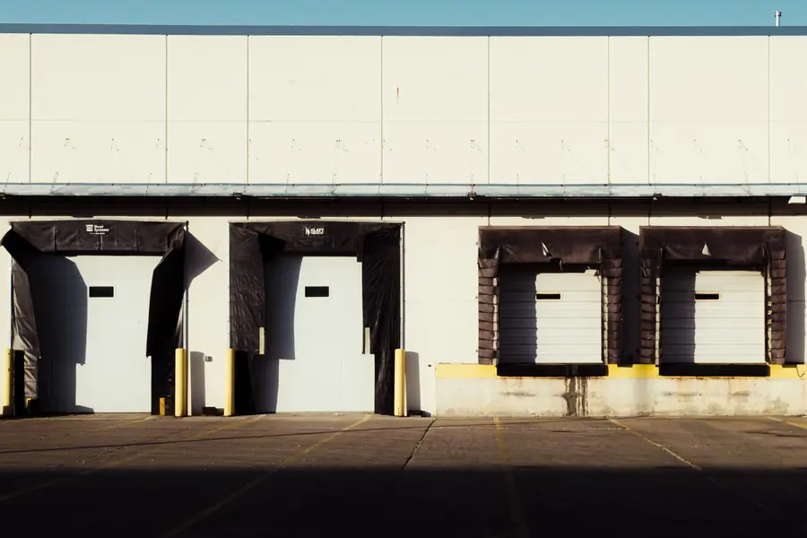 A row of empty loading dock doors with yellow bollards outside a warehouse in bright sun.